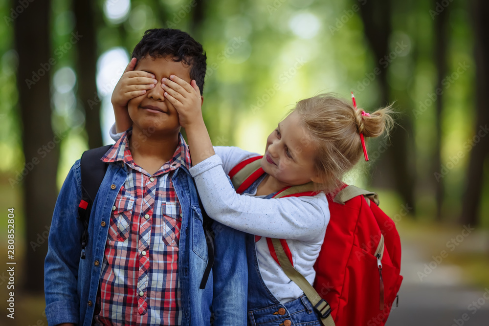 Fototapeta premium Two school Friends having fun in park. Caucasian Girl covering eyes to surprised afro american boy.