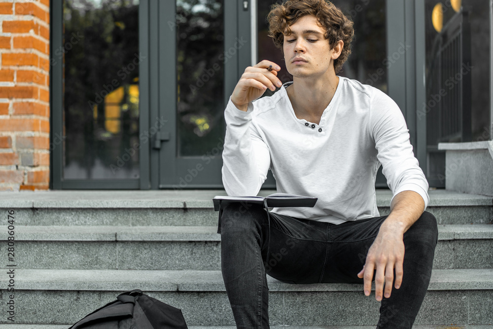 Pensive student sitting on modern building steps and making notes in notepad.