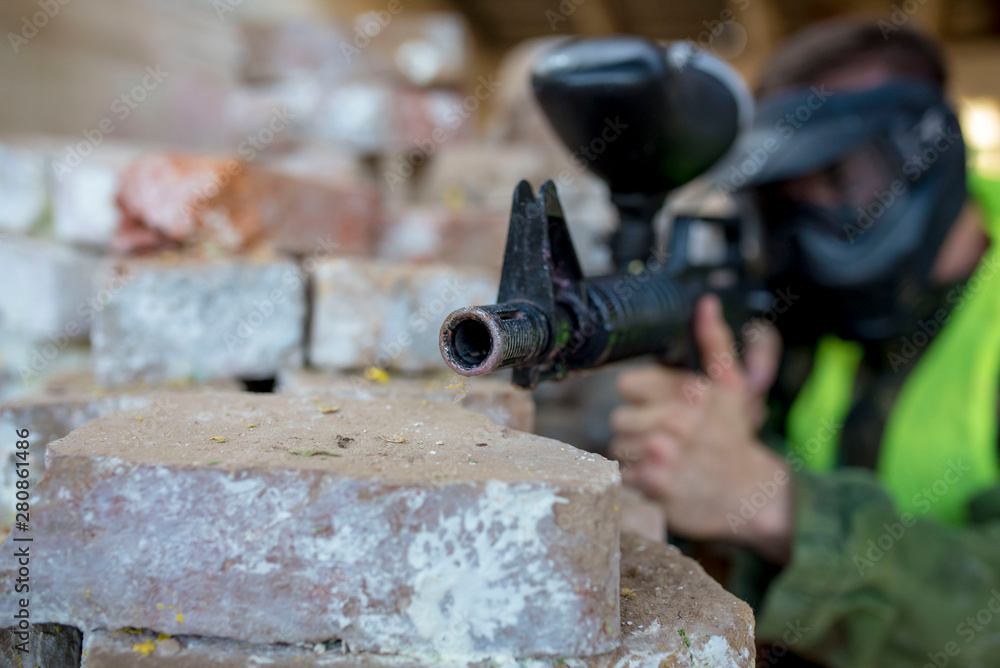 Close up of male paintball player with protective mask and uniform shooting with paintball gun on paintball battlefield 