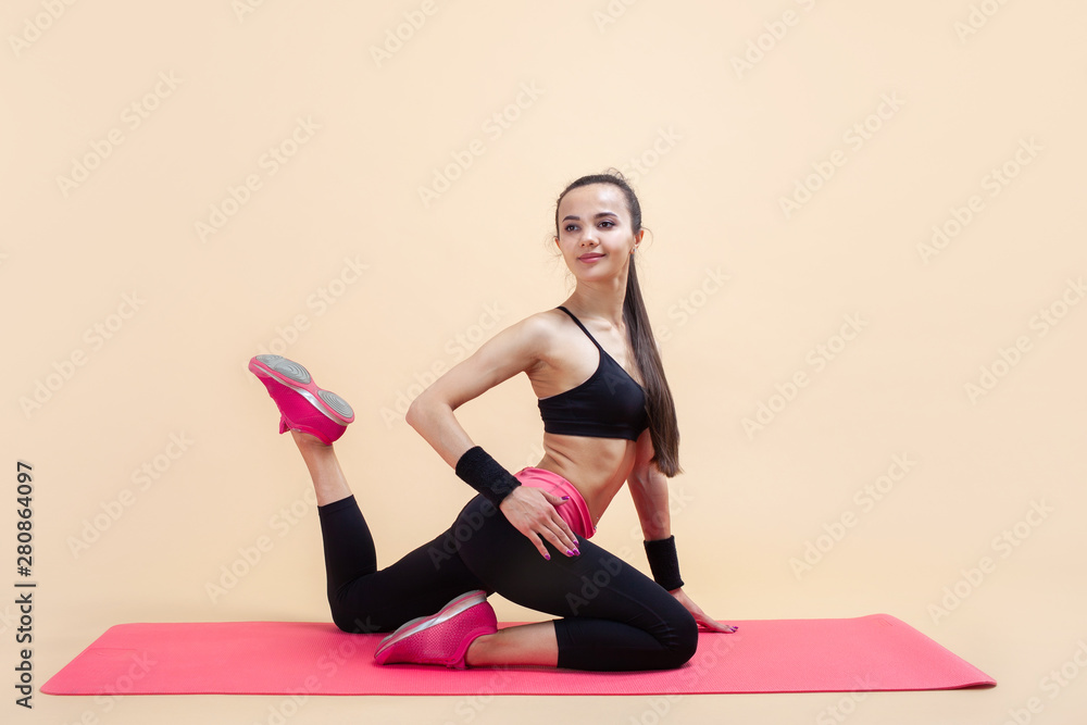 A young attractive girl sits in a pose of a mermaid with a hand on the waist on a pink yoga mat, on a pink background. A young attractive girl sits in a pose of a mermaid with a hand on the waist on a pink yoga mat, on a pink background.
