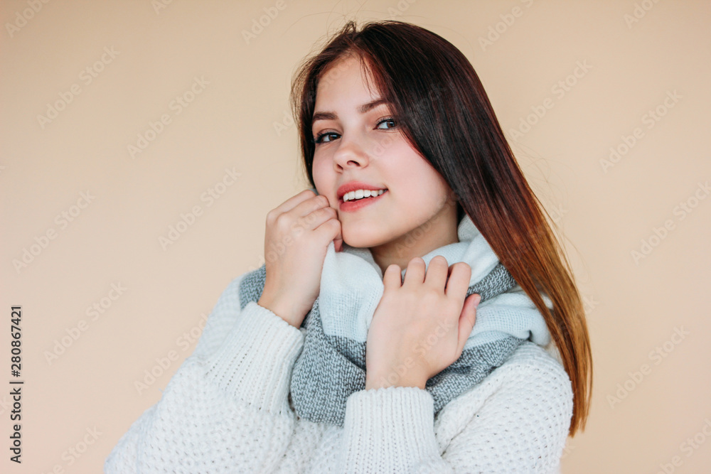 Beautiful smiling girl with clean skin and dark long hair in cozy white sweater and warm scarf on beige background Beautiful smiling girl with clean skin and dark long hair in cozy white sweater and warm scarf on beige background