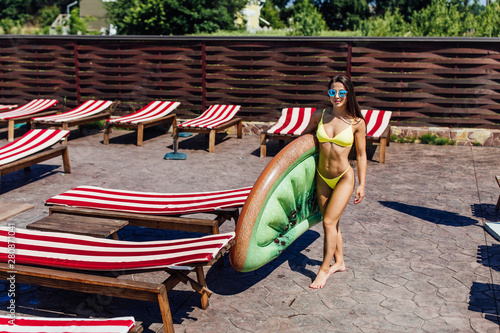 Vacation. Pretty young girl wearing swimsuit standing near pool, holding inflatable mattress kiwi. Weekend. © Тарас Нагирняк
