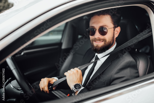 Handsome young smiling bearded driver in full suit with fastening seat belt driving a car. © Тарас Нагирняк