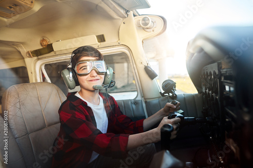 cheerful little boy sitting in cockpit in front of control board with wheel in hands, dreams to be a pilot when he is grown up, looking in the sky with joyful smile.
