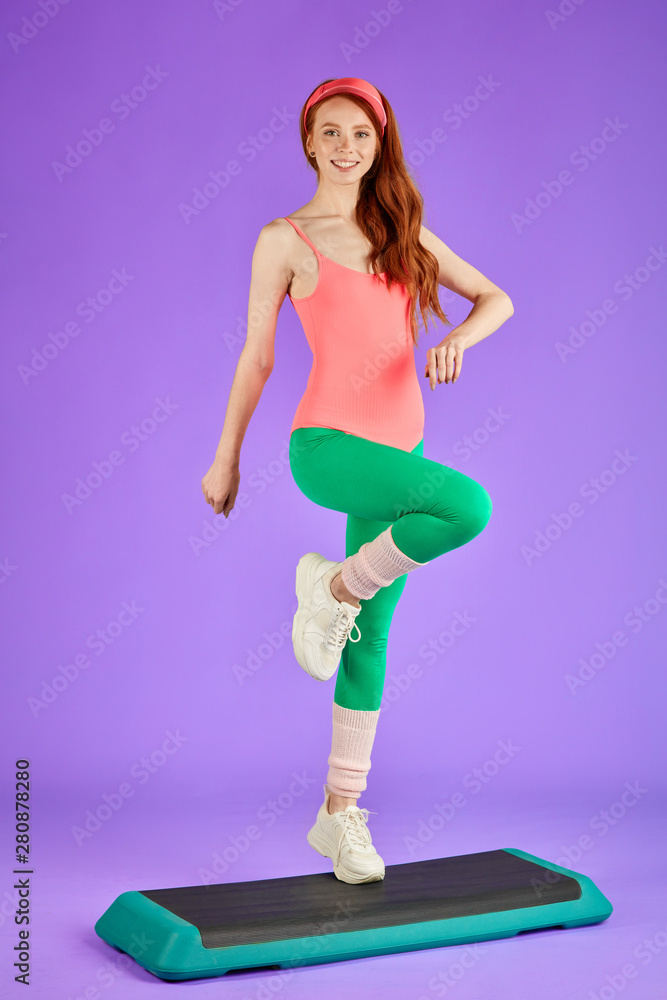 vertical full length view of young pretty fit woman makes intense steps at  fitness platform, prefers home workouts, wants to lose some weight, wears  80s retro style colorful aerobics outfit Photos