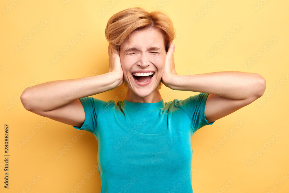Portrait of pretty, unhappy, angry woman in blue T-shirt closes ears with hands isolated on yellow background. close up portrait Portrait of pretty, unhappy, angry woman in blue T-shirt closes ears with hands isolated on yellow background. close up portrait