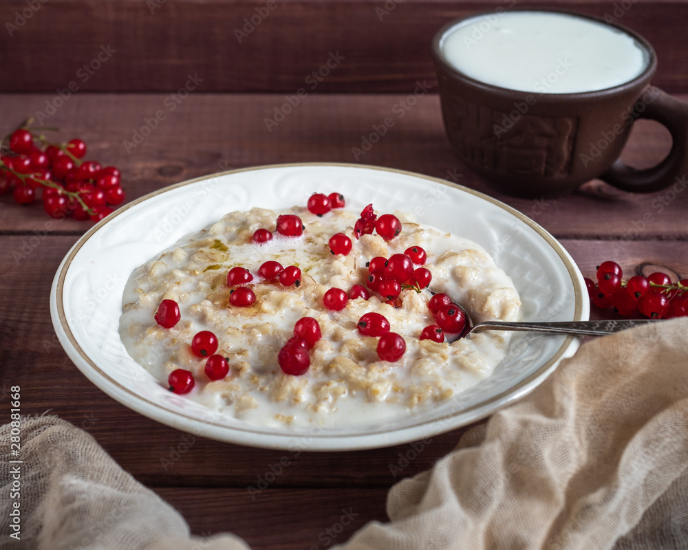 Breakfast of oatmeal milk porridge with fresh berries of currant and milk, a spoon and a mug of milk on a wooden tray Breakfast of oatmeal milk porridge with fresh berries of currant and milk, a spoon and a mug of milk on a wooden tray