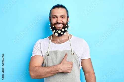 cheerful positive man with beard decorated with flowers ahowing thumb up. close up portrait. good job, body language. isolated blue background photo