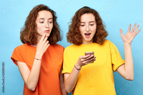 Two girls twins smiling, looking smart phone over blue background.girl received message about winning, looks at screen with sceptic expression finger on lips trying to read sister's chat, sms photo