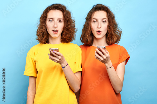 surprised emotional twins with wide open mouth and bugged eyes holding smart phones and looking at the camera. sale, business, free message, double discounts photo