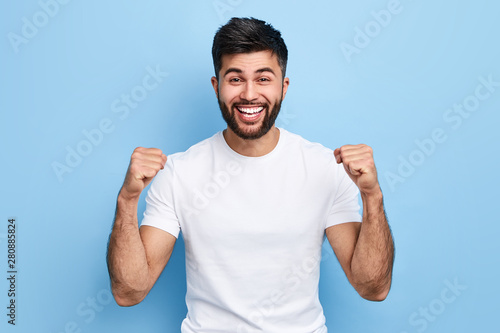 excited Arab man celebrating success with two fists in air isolated on the blue background. close up portrait, studio shot , happiness, positive emotion and feeling. I've done it. facial expression photo