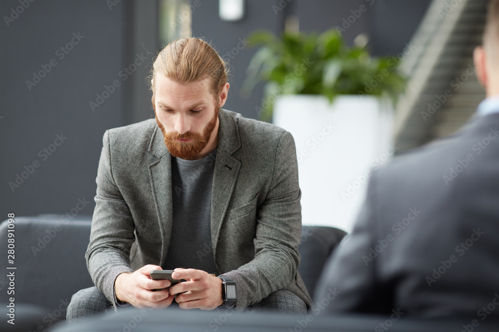 Serious young bearded man in gray jacket sitting in lobby and using internet app on smartphone Serious young bearded man in gray jacket sitting in lobby and using internet app on smartphone