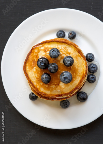 Pancakes with blueberries and honey on a white plate on a black wooden background, overhead view. Flat lay, top view, from above.
