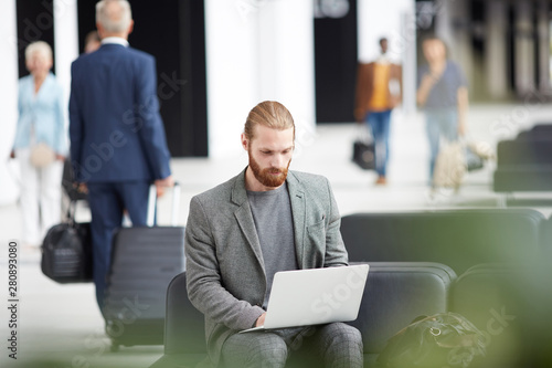 Serious concentrated young man with red beard sitting in airport and typing on laptop