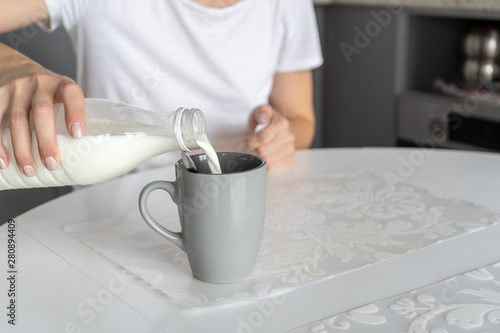 On a white table in the kitchen there is a gray cup with freshly made morning coffee. A woman pours milk from a bottle into a cup.