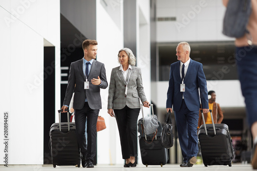 Confident team of forum participants in formalwear carrying wheeled suitcases and crossing airport together of guide