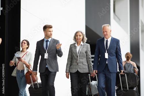 Group of multi-ethnic business people in suits moving along airport with suitcases and listening to young colleague