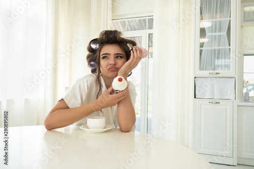 beautiful young brunette woman in hair curlers holding cupcakes in both hands sitting with cup of coffee in bright kitchen looking upset