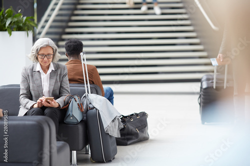 Content elderly Asian lady with gray hair sitting on black leather sofa in airport and browsing internet on smartphone