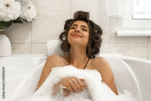 attractive young brunette woman in hair curlers enjoying her bath with foam in bright bathroom photo