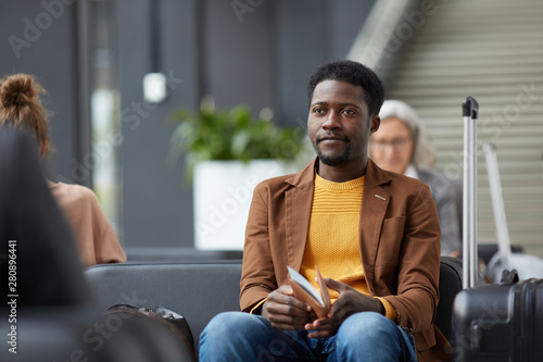 Content Afro-American guy with beard sitting in airport waiting area and holding passport and boarding pass