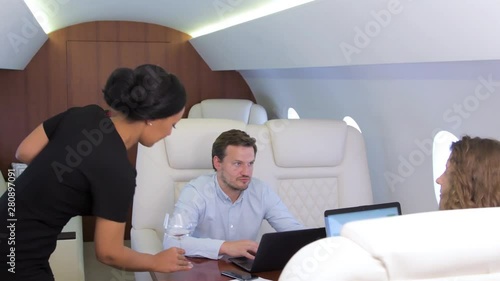 Work on laptop on board of private jet. Biracial flight attendant offering and pouring glass of wine for caucasian businessman and businesswoman travel inside of business airplane cabin. photo