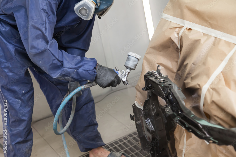A male worker in jumpsuit and gloves paints with a spray gun a front frame part of the car body in black after being damaged at an accident. Auto service industry professions A male worker in jumpsuit and gloves paints with a spray gun a front frame part of the car body in black after being damaged at an accident. Auto service industry professions