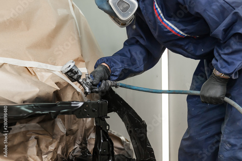 A male worker in jumpsuit and gloves paints with a spray gun a front frame part of the car body in black after being damaged at an accident. Auto service industry professions
