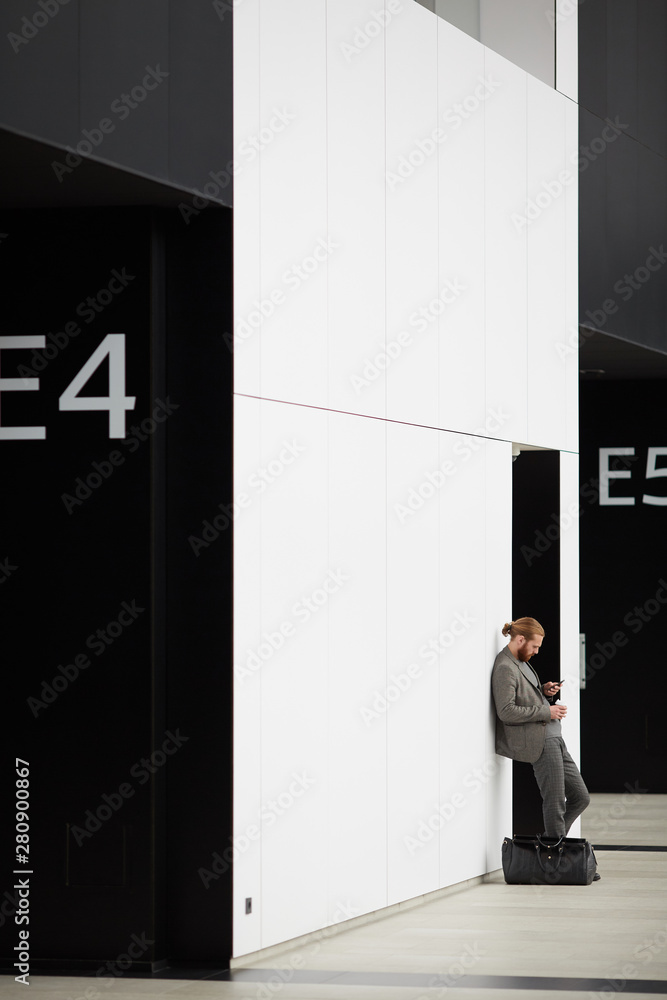 Fototapeta premium Young hipster man with ponytail and beard leaning on wall and using wifi on gadget while spending time in airport