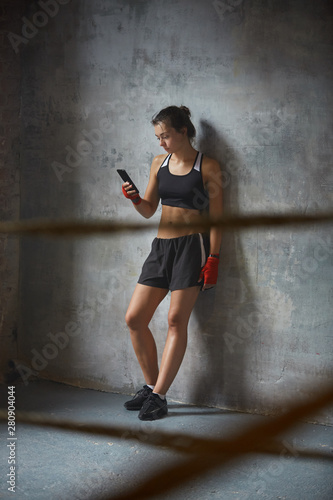 Full length portrait of young athletic woman using smartphone leaning on concrete wall, copy space