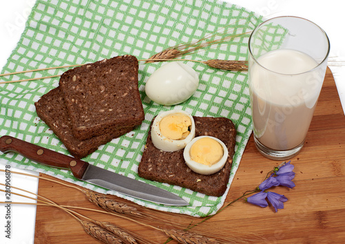 Slices of bread with eggs, glass of milk, rye and bluebell flower on checkered table-napkin