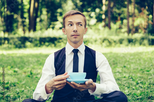 Young attractive man in a vest and tie  resting on the nature and drinking coffee. Holds a cup of tea.