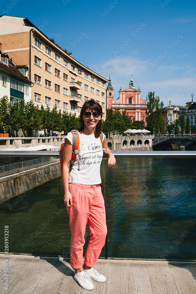 Young smiling woman tourist standing on a bridge in the old centre of Ljubljana, Slovenia, wearing white shirt, white sneakers and coral trousers. With an orange backpack. Travel concept.