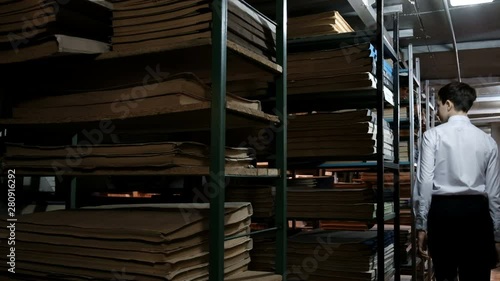 A teenager in a white shirt is looking for a little boy between the shelves with old books. Dark room in the library or archive room with shelves of old literature and documents.