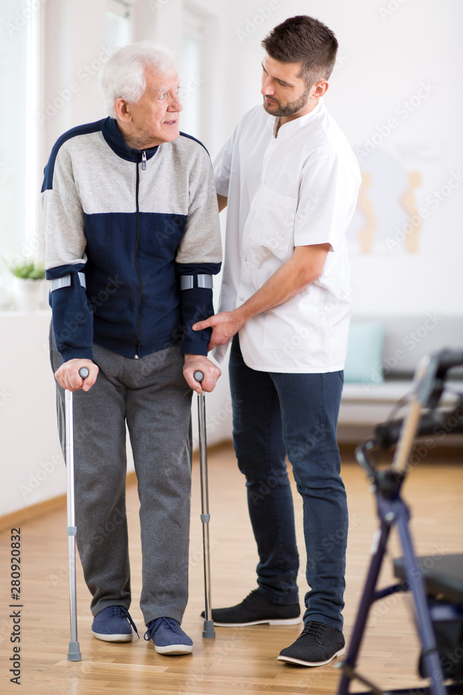 Elderly man walking on crutches and a helpful male nurse supporting him Elderly man walking on crutches and a helpful male nurse supporting him