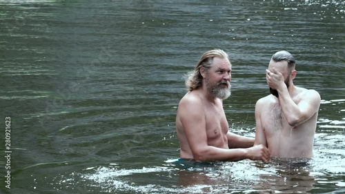 A grown man with a beard, like a priest, dips a young bearded guy into the water. The concept of baptism in the river or entertainment on vacation. People and nature, religion and lifestyle.