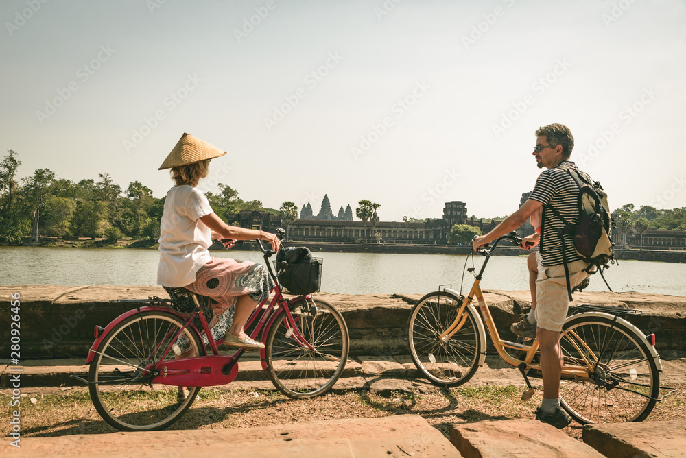Fototapeta premium Tourist couple cycling in Angkor temple, Cambodia. Angkor Wat main facade reflected on water pond. Eco friendly tourism traveling. Toned