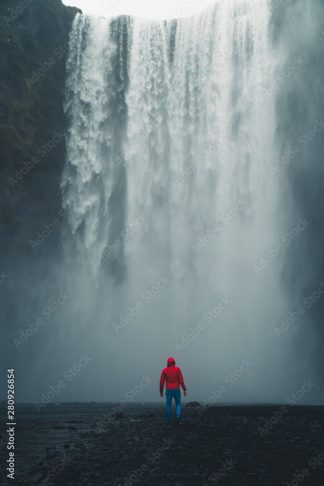 Fototapeta premium Skogafoss waterfall Iceland. Man standing against huge waterfall surrounded by green hills. Spring in Iceland.