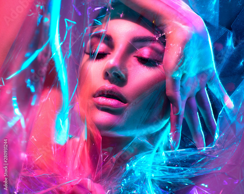 Fashion model woman in colorful bright neon lights posing in studio through transparent film. Portrait of beautiful sexy girl in UV. Art design colorful makeup