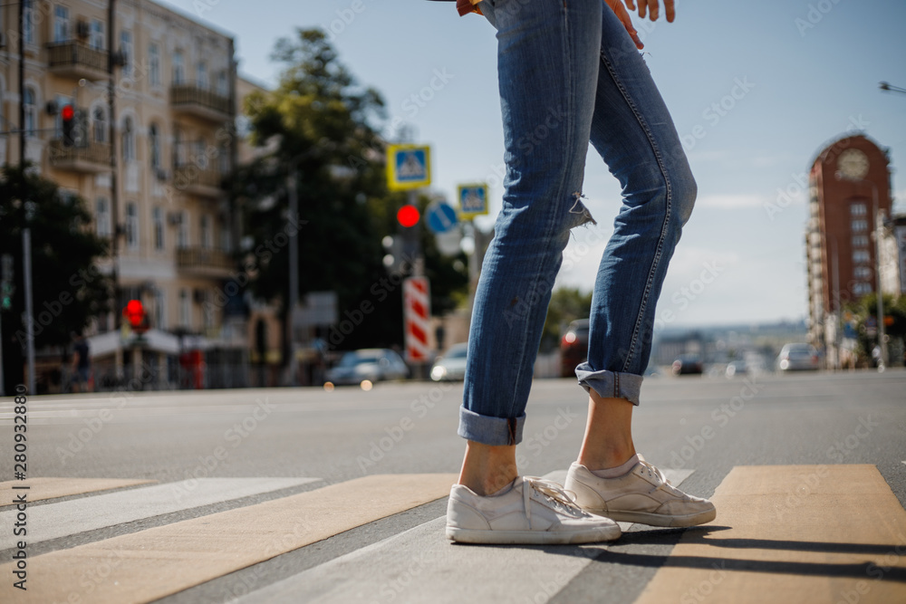 Young girl dressed in jeans and sneakers is crossing the road with a backpack in a city street on a summer sunny day Young girl dressed in jeans and sneakers is crossing the road with a backpack in a city street on a summer sunny day