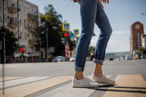 Young girl dressed in jeans and sneakers is crossing the road with a backpack in a city street on a summer sunny day