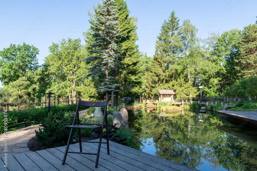 Dawn. A folding wooden chair stands on a deck of wooden boards in front of a small picturesque pond