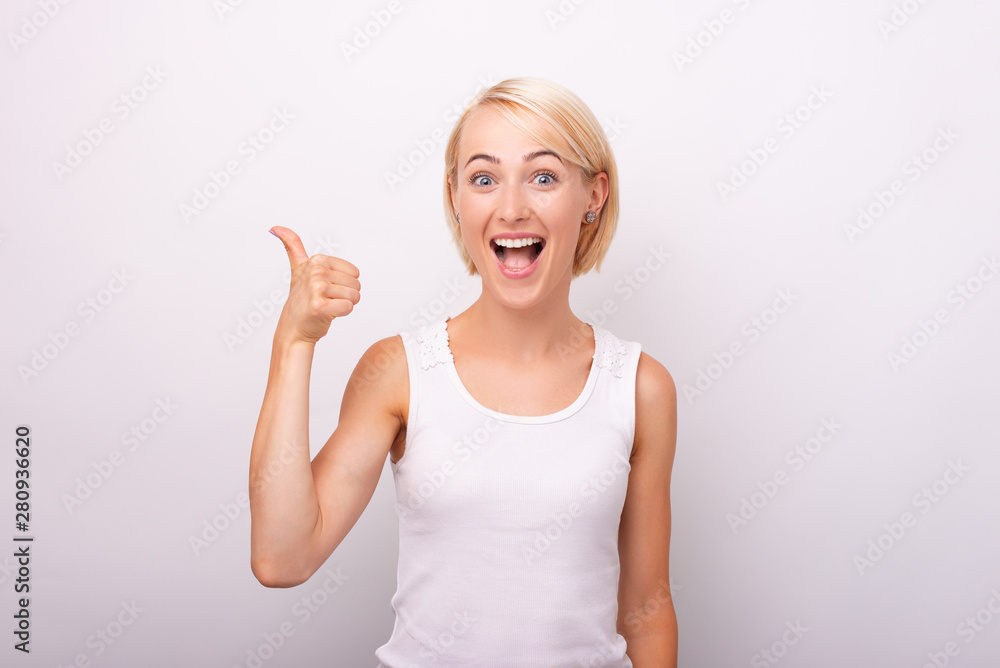 Portrait of beautiful amazed woman showing thumbs up gesture over white wall Portrait of beautiful amazed woman showing thumbs up gesture over white wall