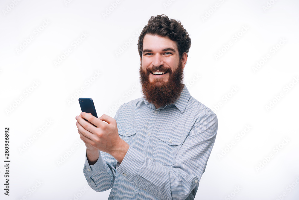 Photo of handsome cheerful young man with beard holding smartphone and looking at the camera over white wall Photo of handsome cheerful young man with beard holding smartphone and looking at the camera over white wall