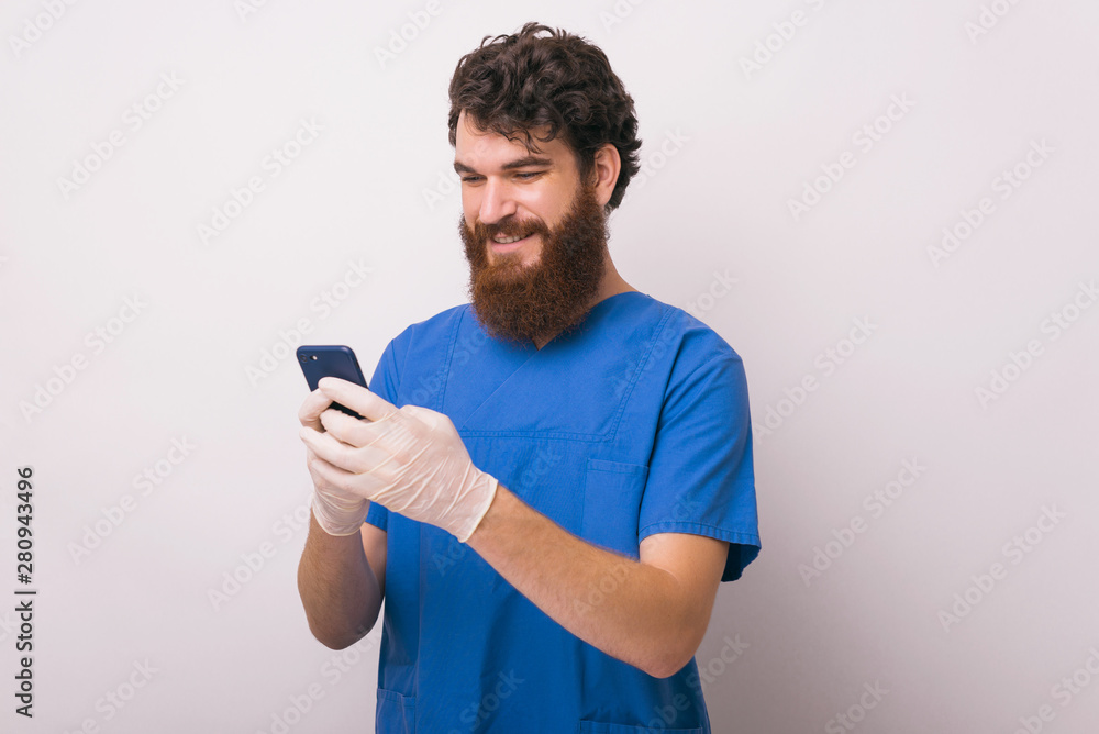 Young smiling doctor looking at phone while standing over white wall Young smiling doctor looking at phone while standing over white wall