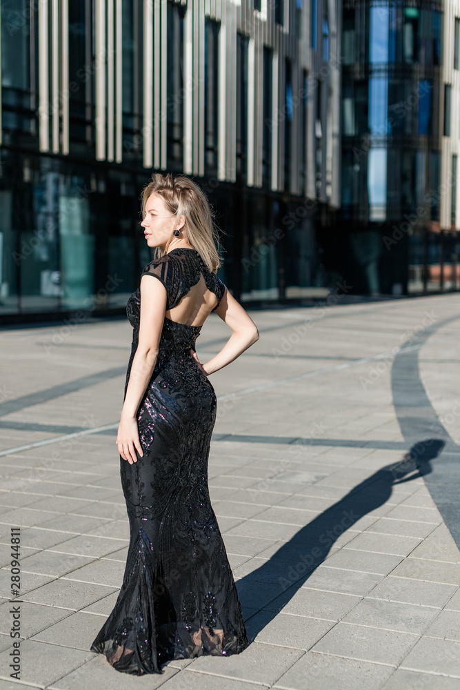 Fototapeta premium A beautiful blonde girl stands with her back in a black evening long dress against the backdrop of a modern building.