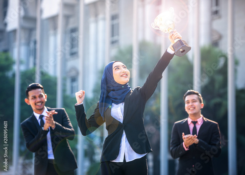 Successful Muslim business woman with arms up holding a trophy and celebrating, with teammates supporting behing. Female Empowerment concept