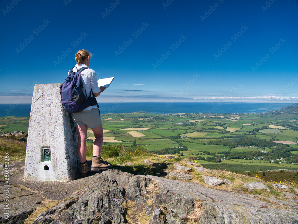 Obraz premium A lone woman walker at the top of Garn Fadryn checks a map while facing Nefyn on the Llyn Peninsula, Gwynedd, Wales, UK