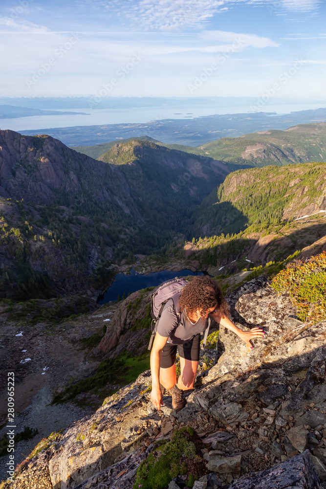 Adventurous girl climbing up a steep cliff in the Canadian Mountain Landscape during a vibrant summer evening. Taken at Mt Arrowsmith, near Nanaimo, Vancouver Island, BC, Canada.