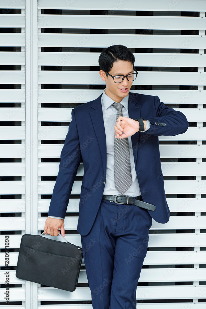Successful young Asian businessman in glasses and classic suit checking time on his watch Successful young Asian businessman in glasses and classic suit checking time on his watch